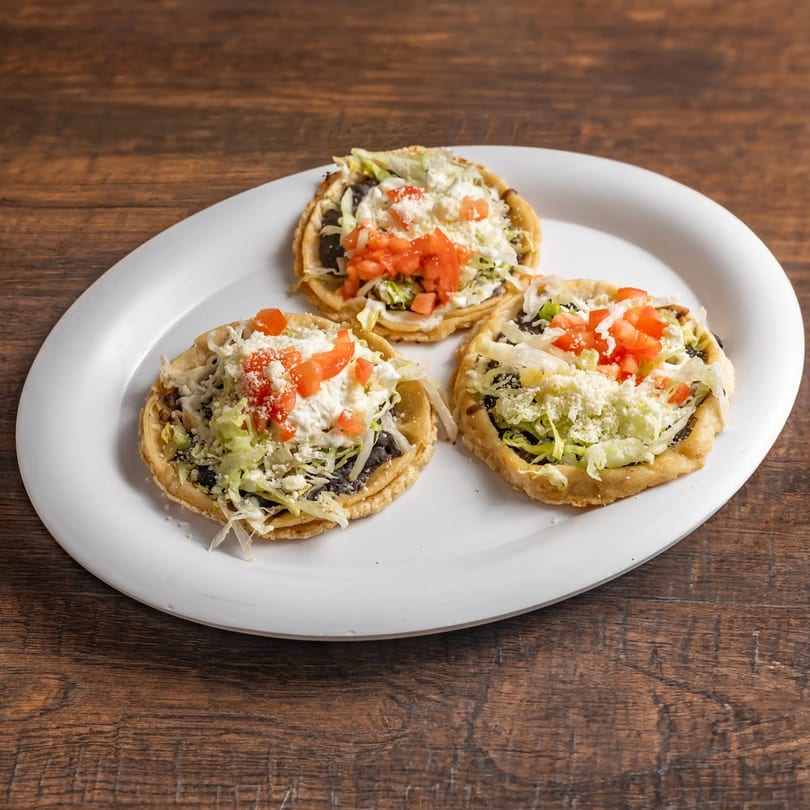 Three Mexican sopes topped with lettuce, tomatoes, cheese, and black beans on a white plate.