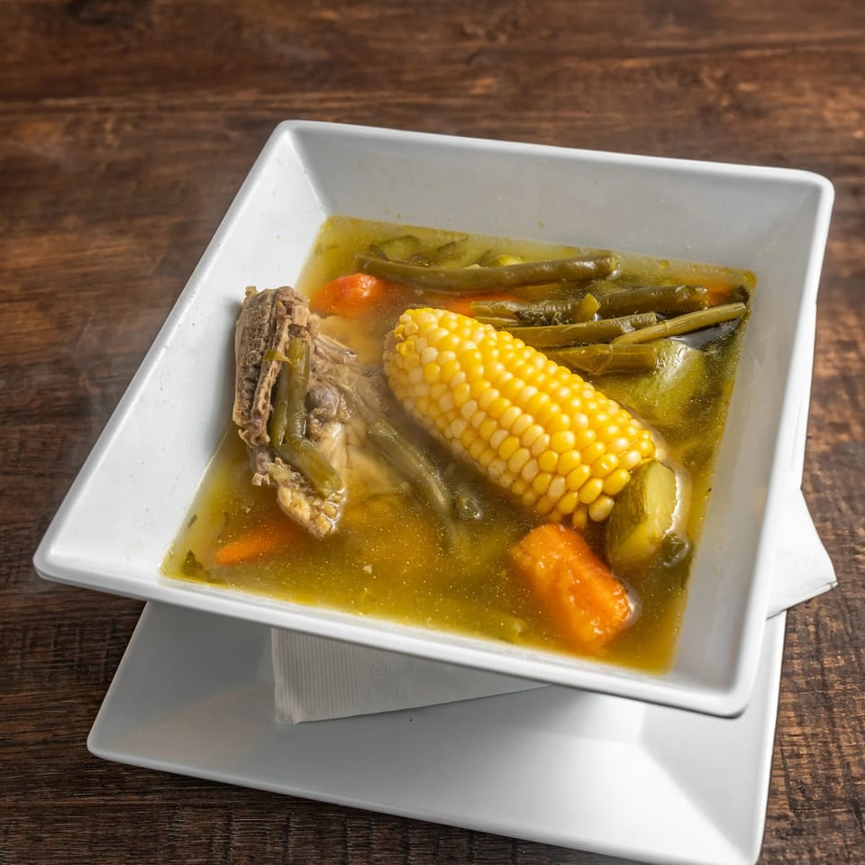 Steaming Mexican soup with corn on the cob, green beans, carrots, and meat in a white bowl.