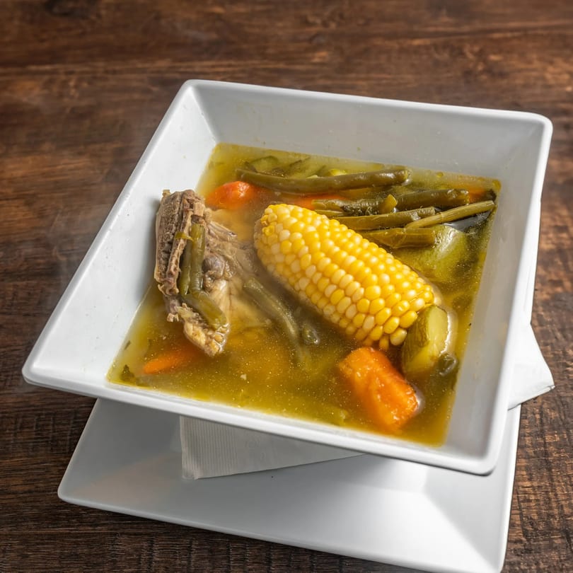 Steaming Mexican soup with corn on the cob, green beans, carrots, and meat in a white bowl.