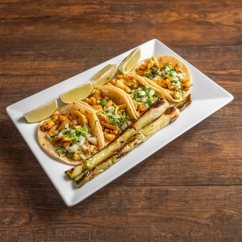 Three chicken tacos with onions, cilantro, lime wedges, and grilled green onions on a white plate.