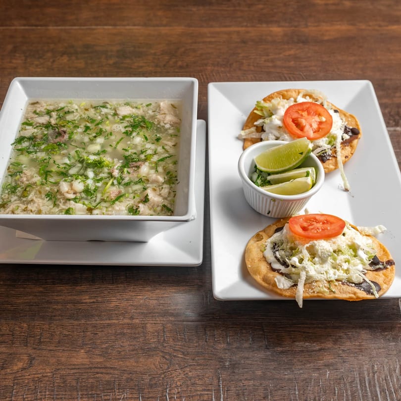 Mexican pozole soup with two sopes topped with cheese, lettuce, tomato, and lime wedges.